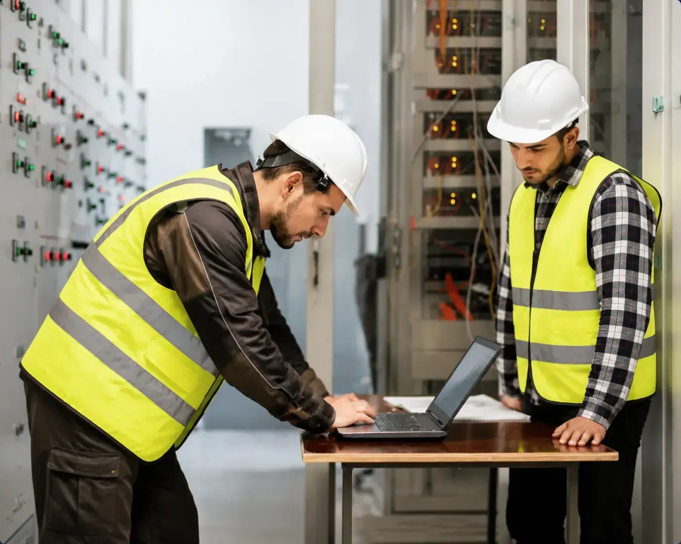 Two technicians in yellow hardhats working on electrical equipment, one using a laptop.