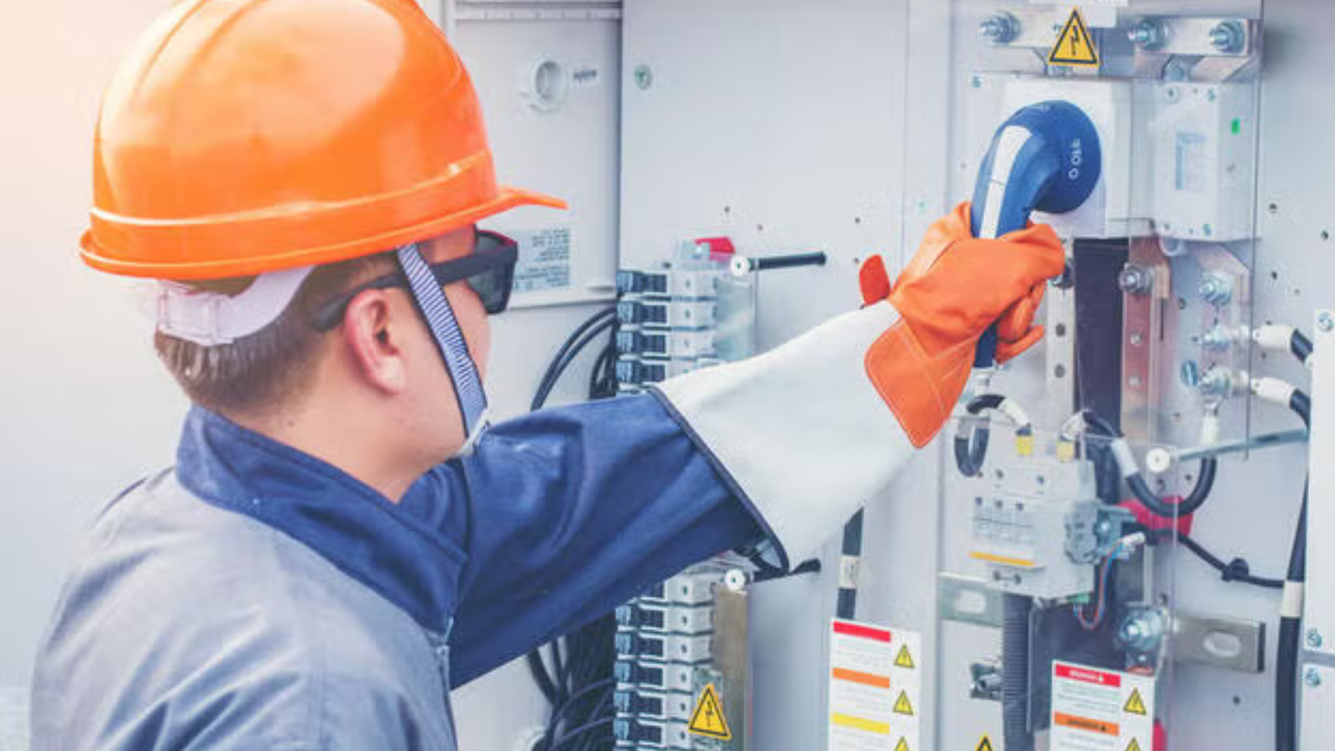 A worker in a hard hat and protective gloves operates electrical equipment inside a control panel.