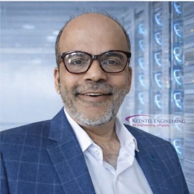 A smiling man with glasses and a beard wearing a blue blazer stands in front of server racks in a data center.