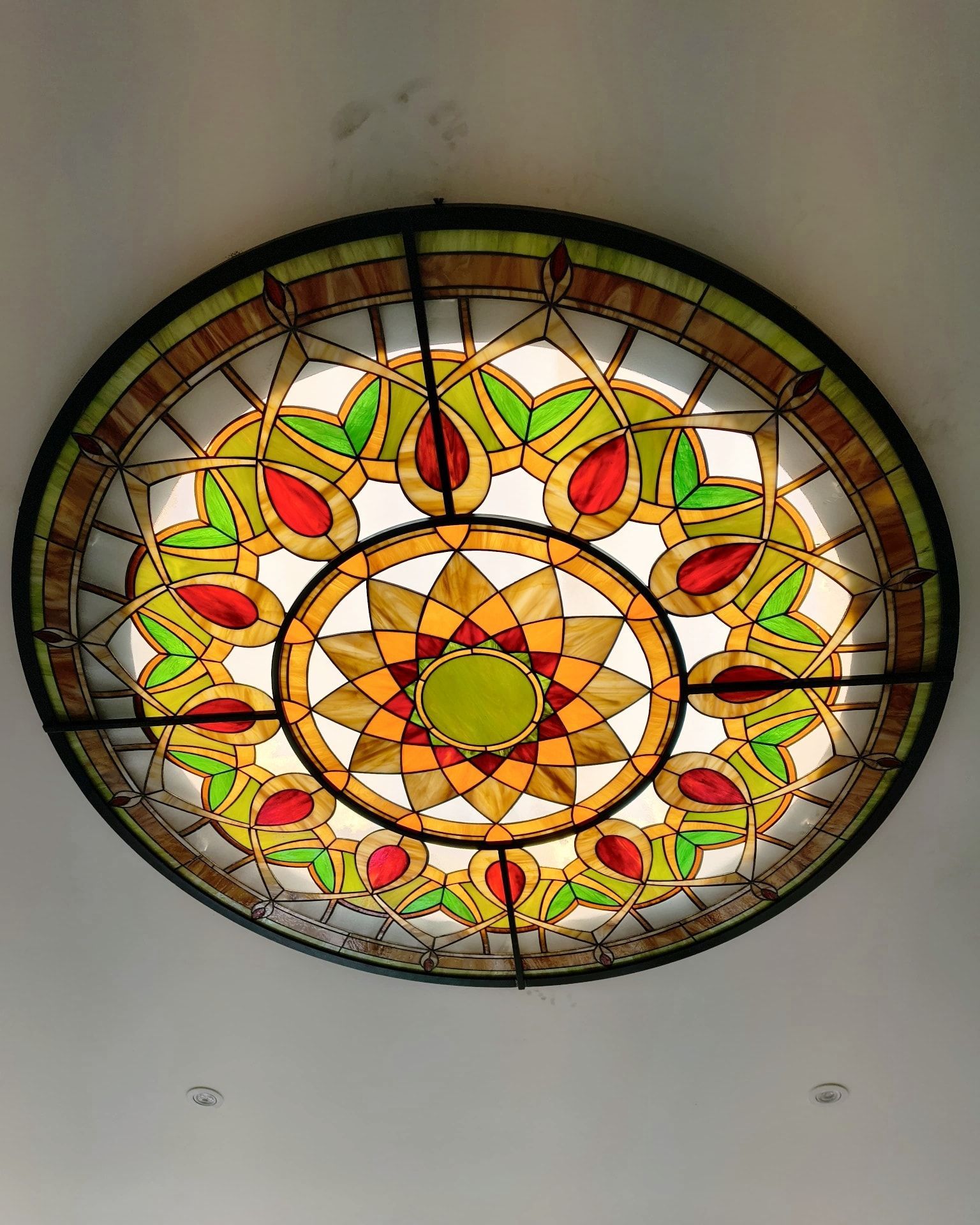 Una lámpara de techo redonda de vidrieras con motivos geométricos en verde, rojo, amarillo y ámbar sobre un techo blanco.