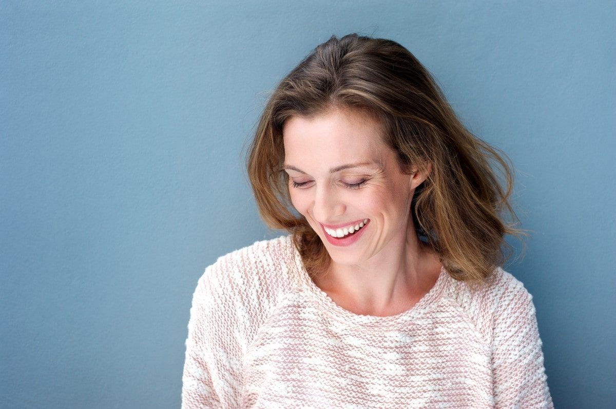 Woman in off-white sweater smiles, looking down against a blue background.