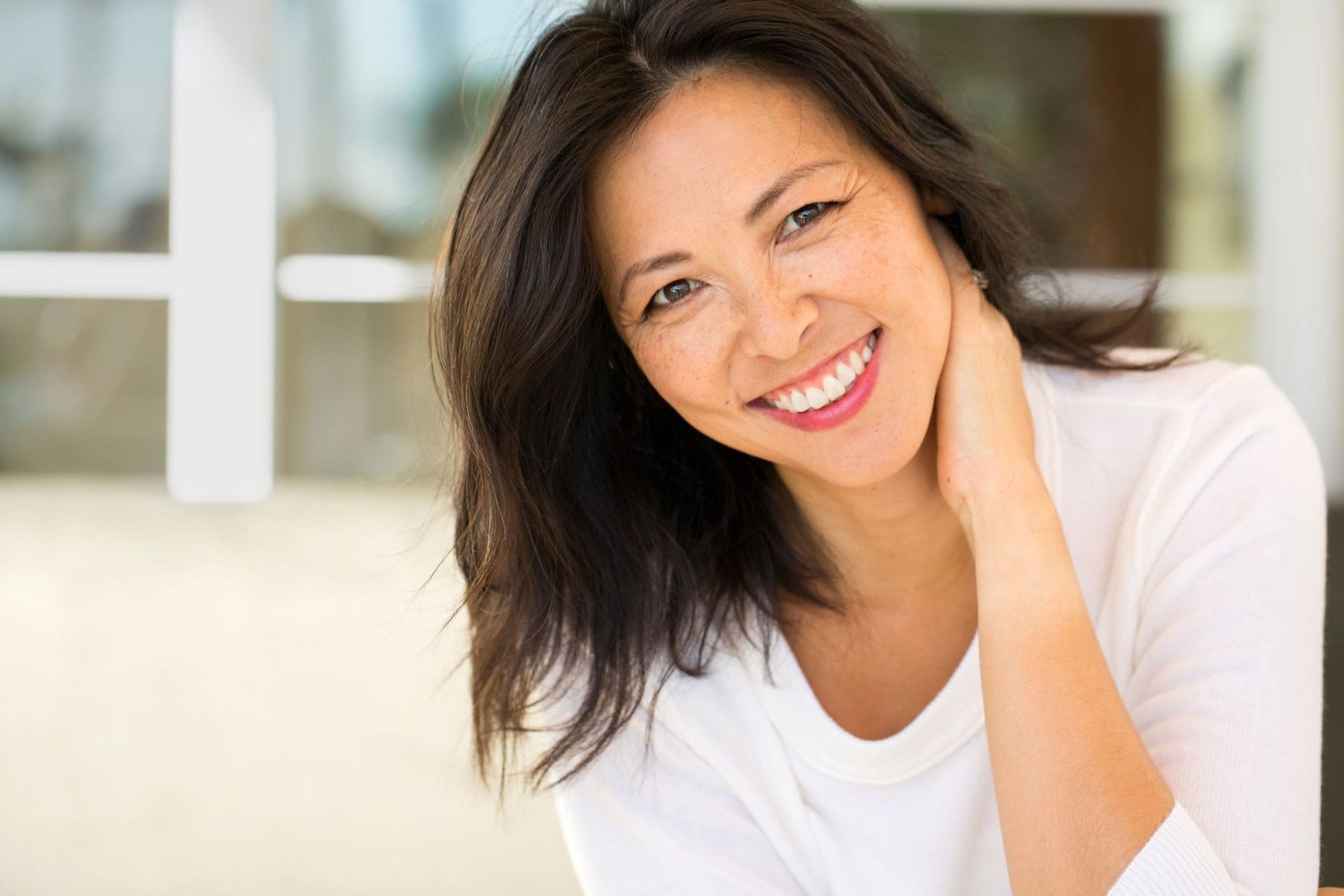 Woman smiling, touching her neck. Sunlight through window in the background.