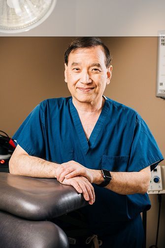 Doctor in blue scrubs smiles, leans on a medical table in a clinic setting.