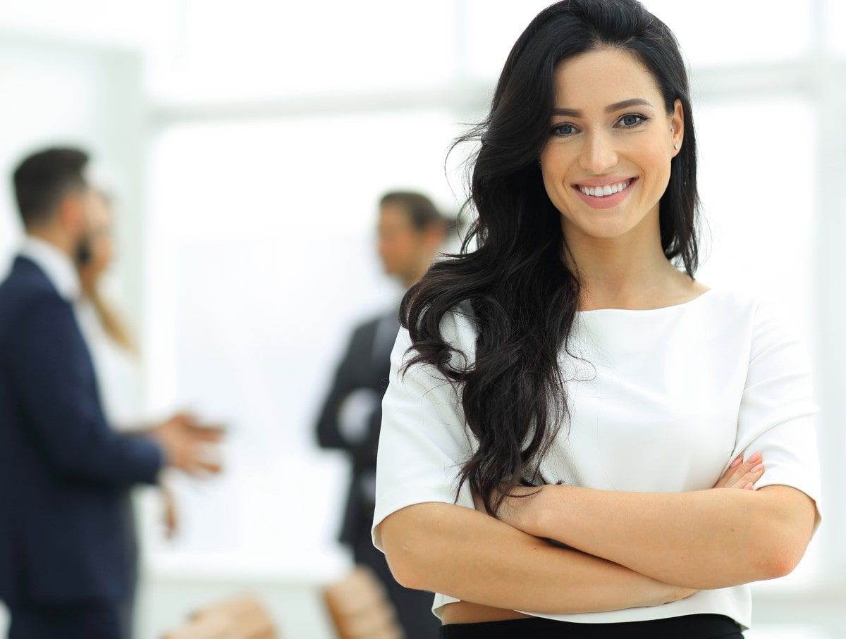 Smiling woman with crossed arms in a business setting, other blurred figures in background.