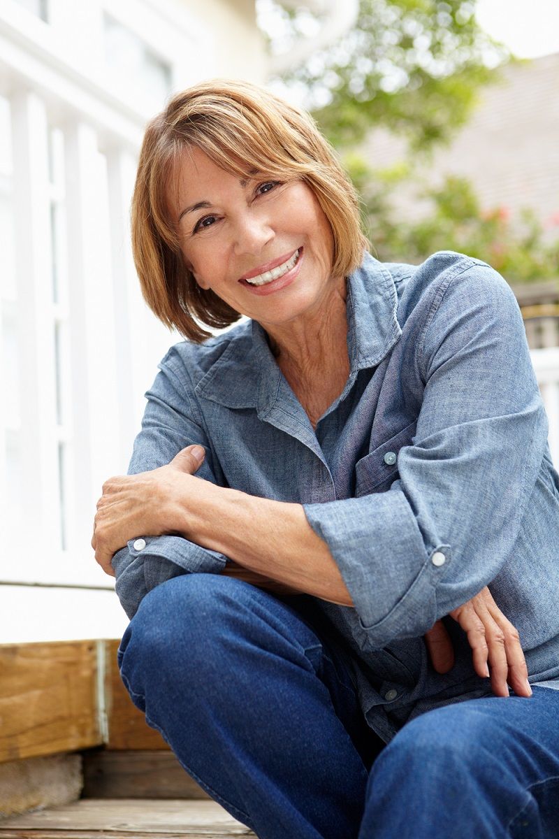 Woman smiling, seated outdoors on wooden steps, wearing denim shirt and jeans.