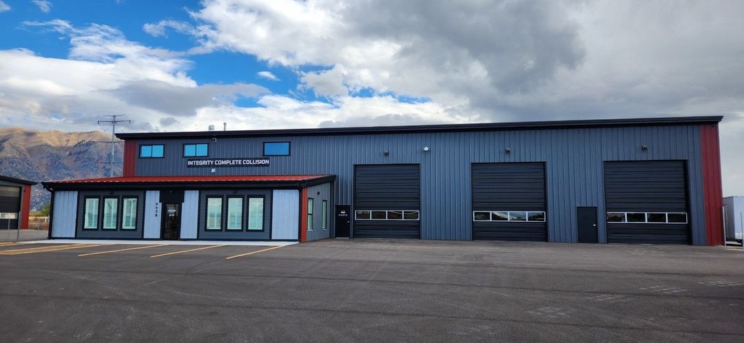Exterior of a metal building with garage doors, office windows, and a mountain backdrop under a cloudy sky.