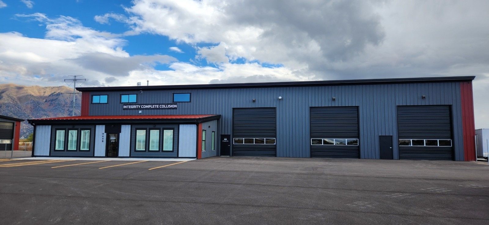 Exterior of a metal building with garage doors, office windows, and a mountain backdrop under a cloudy sky.