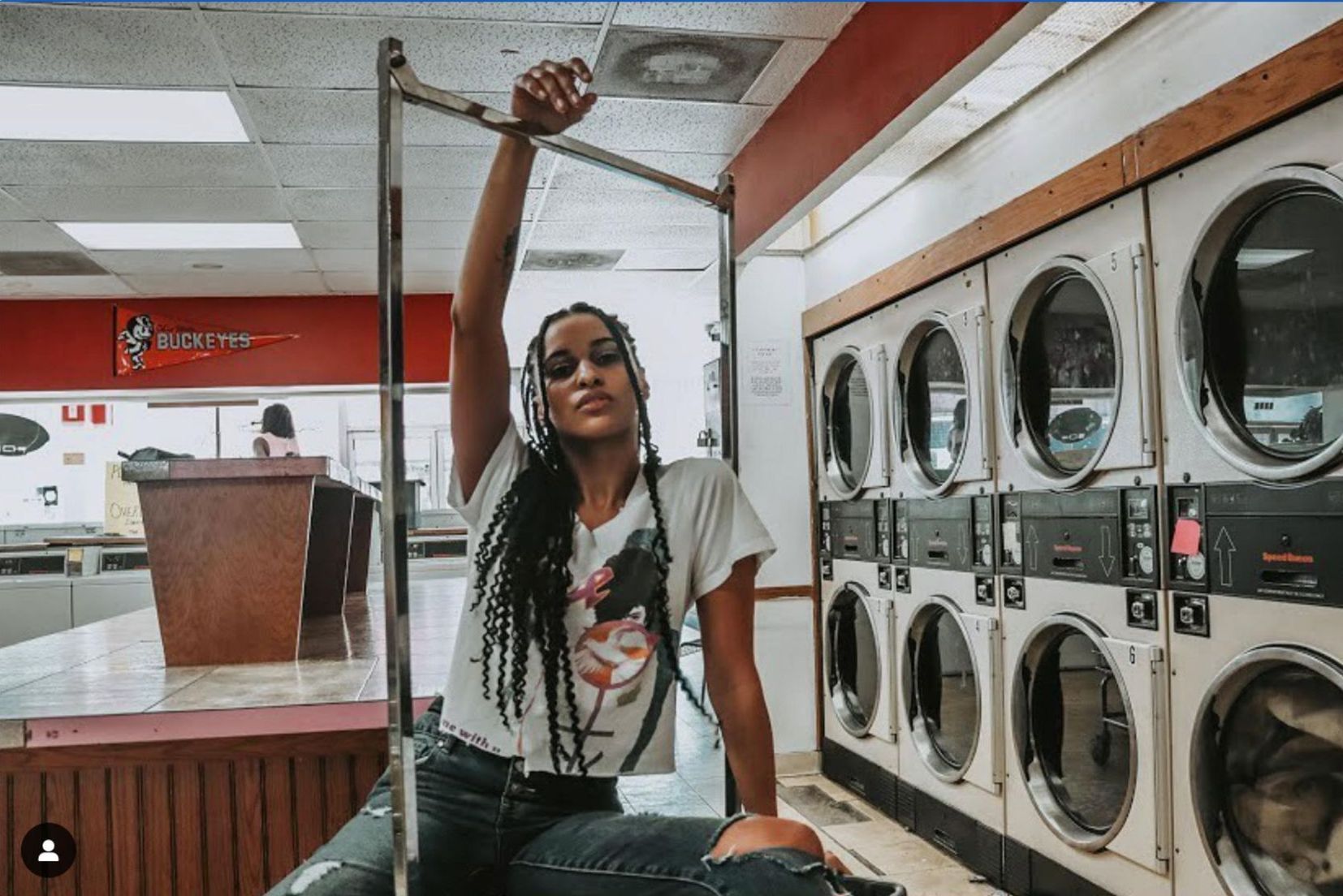 A woman is sitting on a pole in a laundromat.