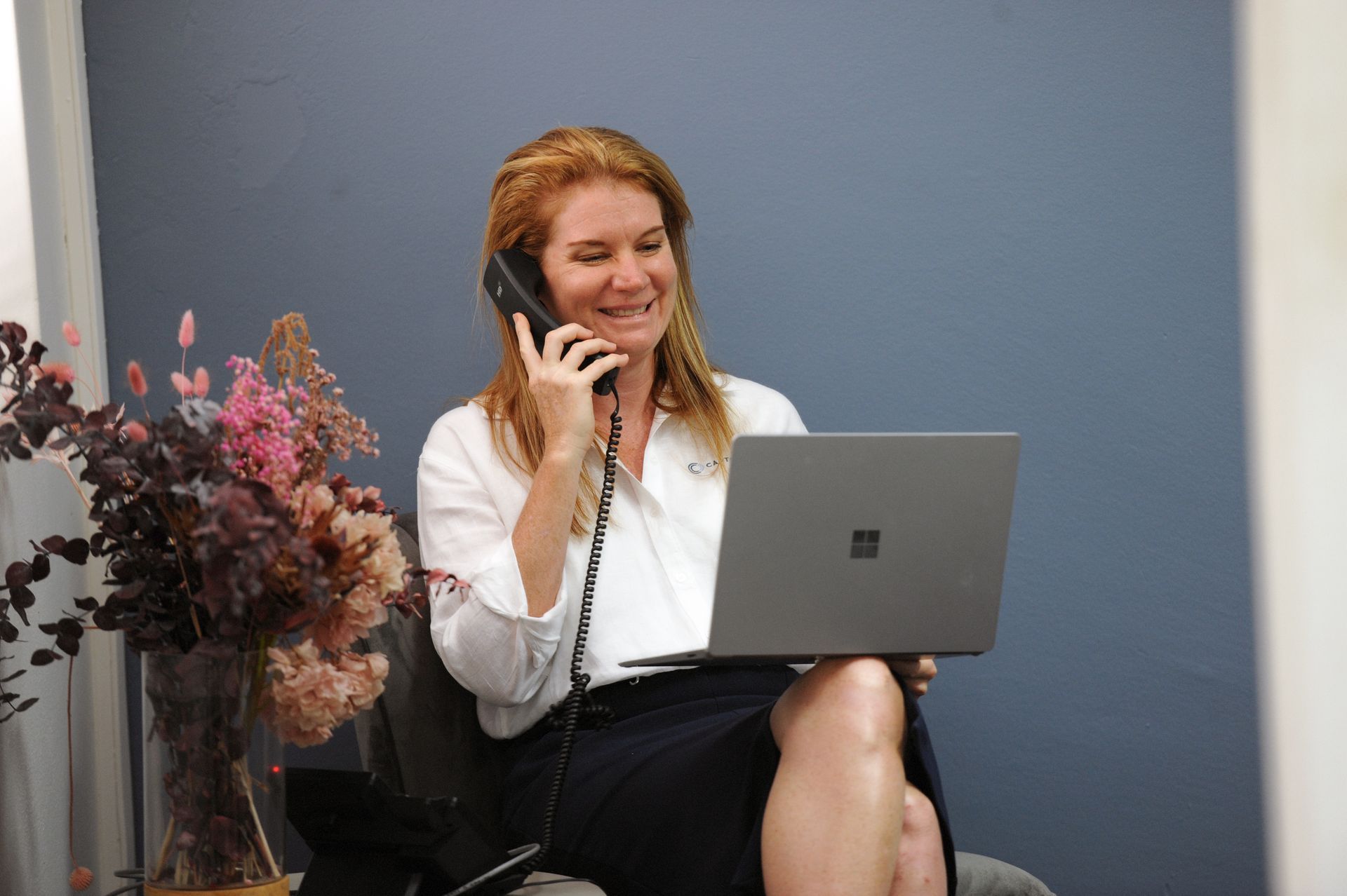 A woman is sitting in a chair talking on a phone while using a laptop computer.