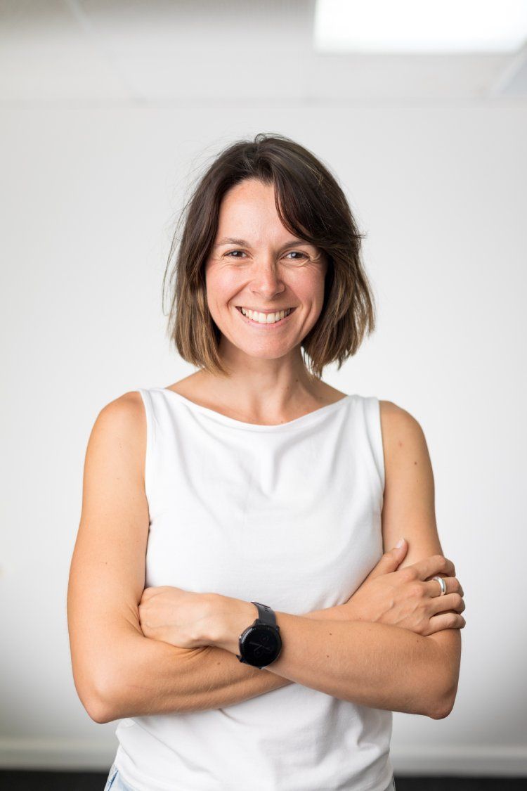 A woman in a white tank top is smiling with her arms crossed.