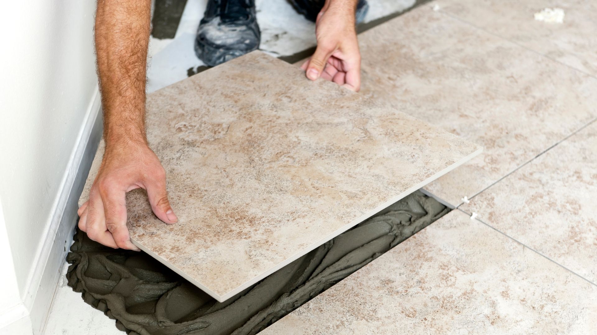Person installing beige floor tile; hands placing tile on wet adhesive.