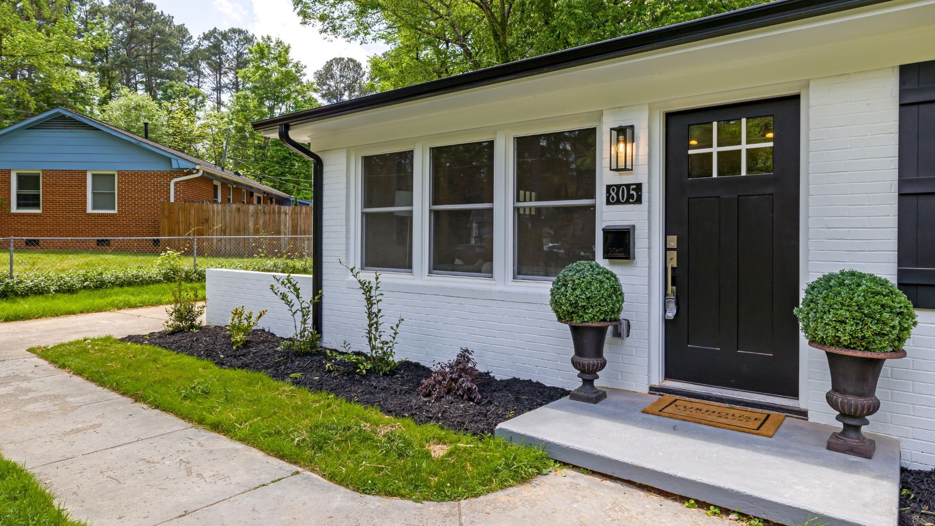 White brick house with black door, shutters, and planters. A small sidewalk and grass lawn.