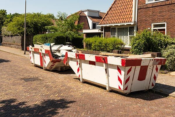 Two dumpsters are parked on the side of the road in front of a house.