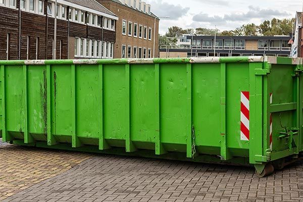 A large green dumpster is parked on a brick driveway in front of a building.