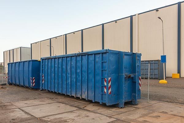 A row of blue dumpsters are parked in front of a building.