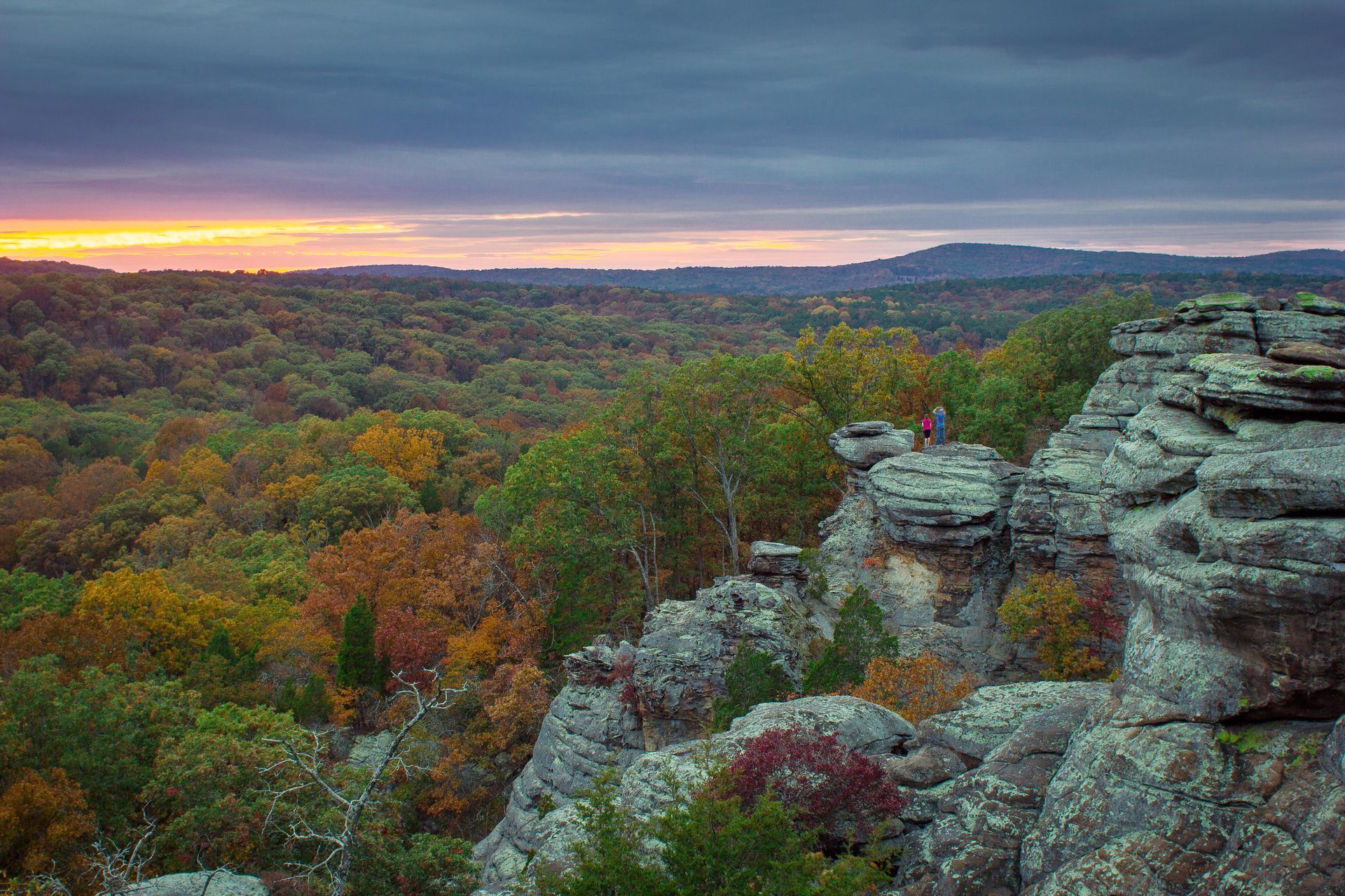 A person is standing on top of a rocky cliff overlooking a forest at sunset.