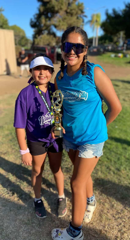 A person wearing a hat and orange shirt, holding a baseball bat on their shoulder on a baseball field.