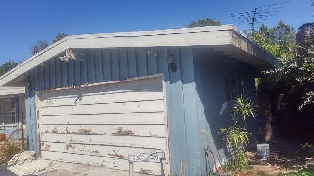 A blue and white garage with a roof is in the backyard of a house.
