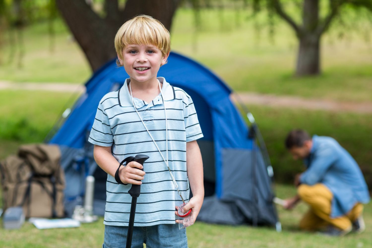 Boy with blonde hair smiles, holding a trekking pole in front of a blue tent in a park.