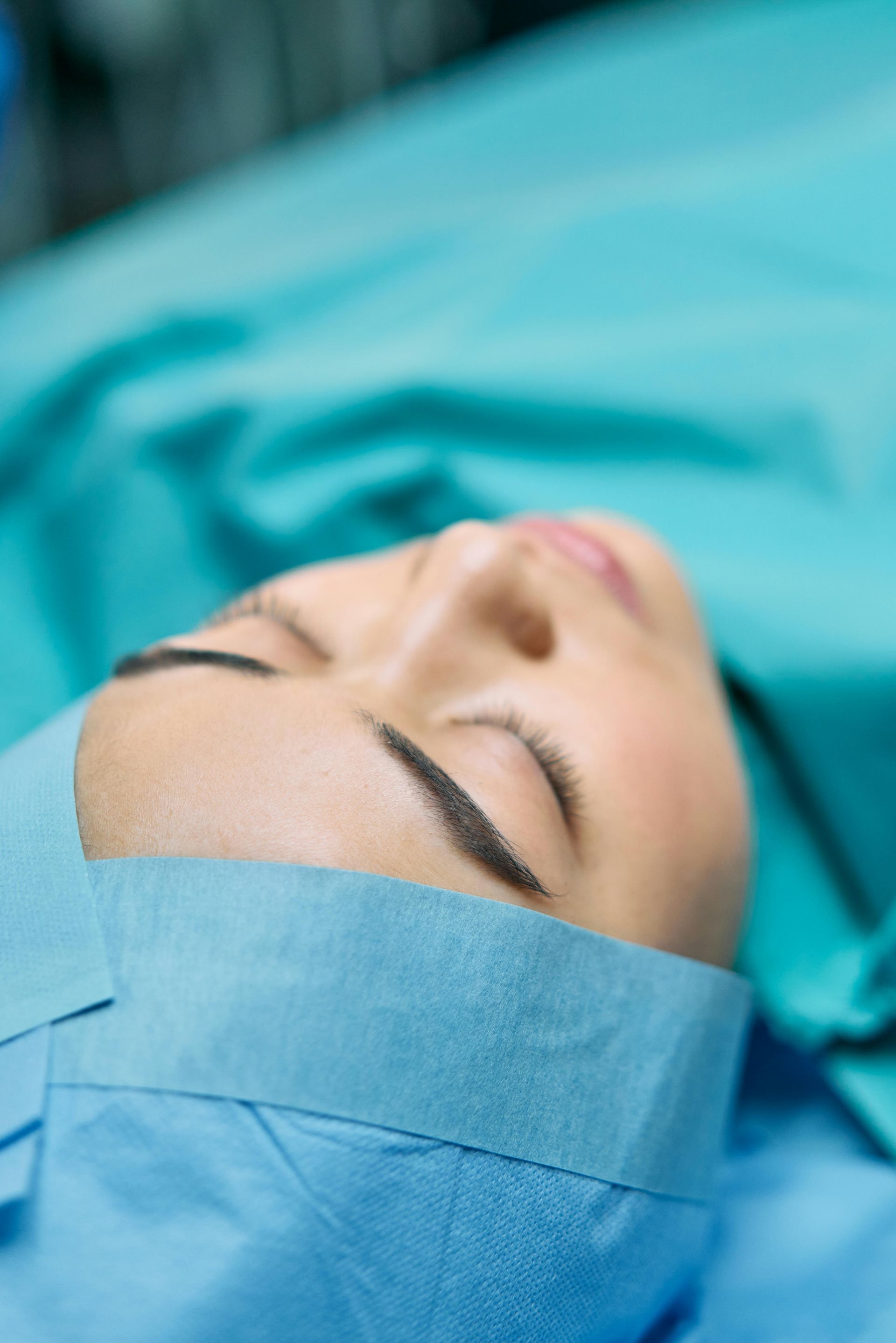 A person lies supine in a hospital, wearing blue surgical headwear and a blue gown against a matching background.