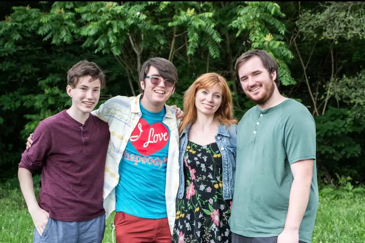 Four people, arms around each other, smile at the camera in front of green trees.