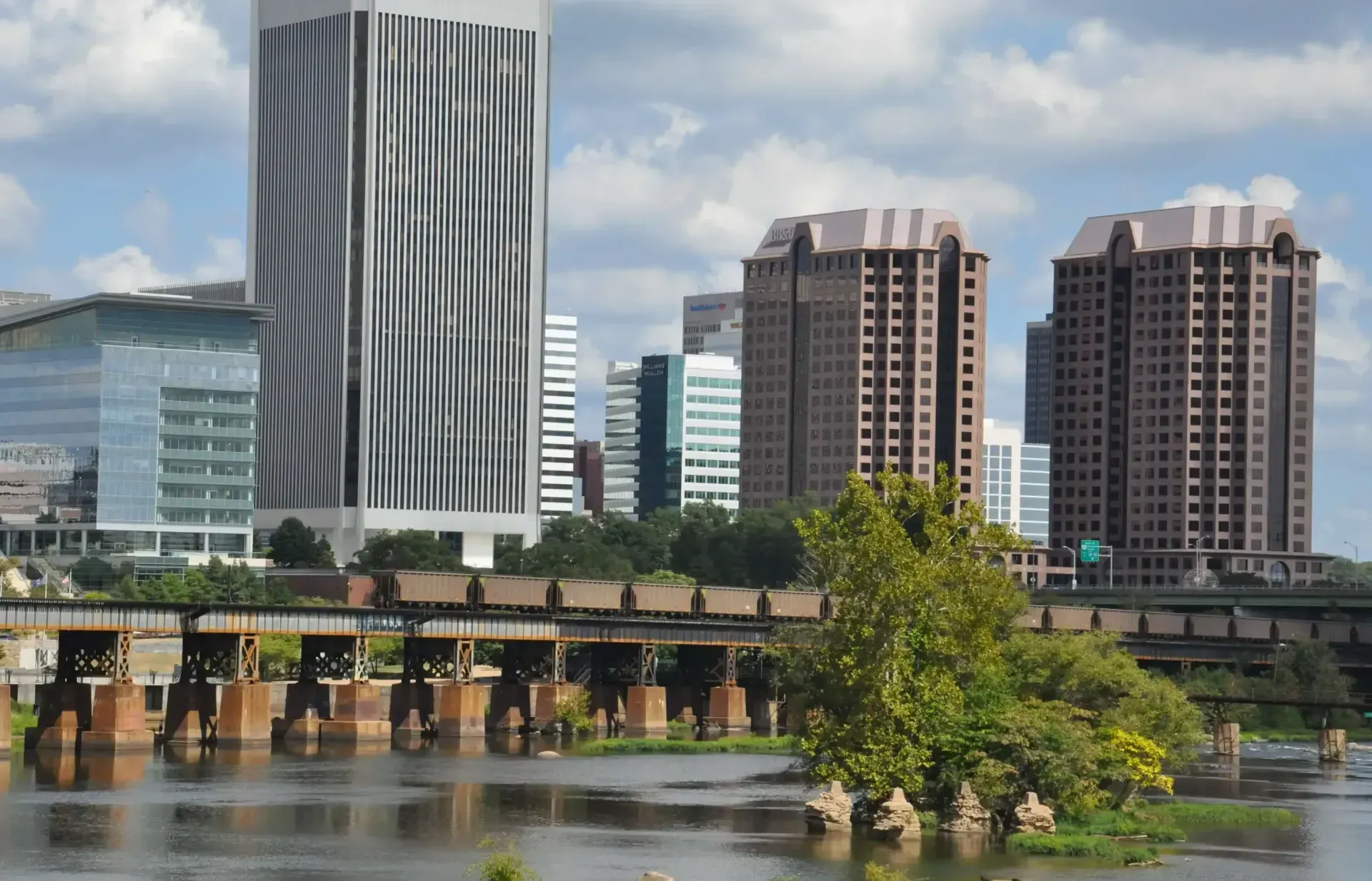 Richmond, Virginia skyline with river and railway bridge in foreground; tall buildings and blue sky.