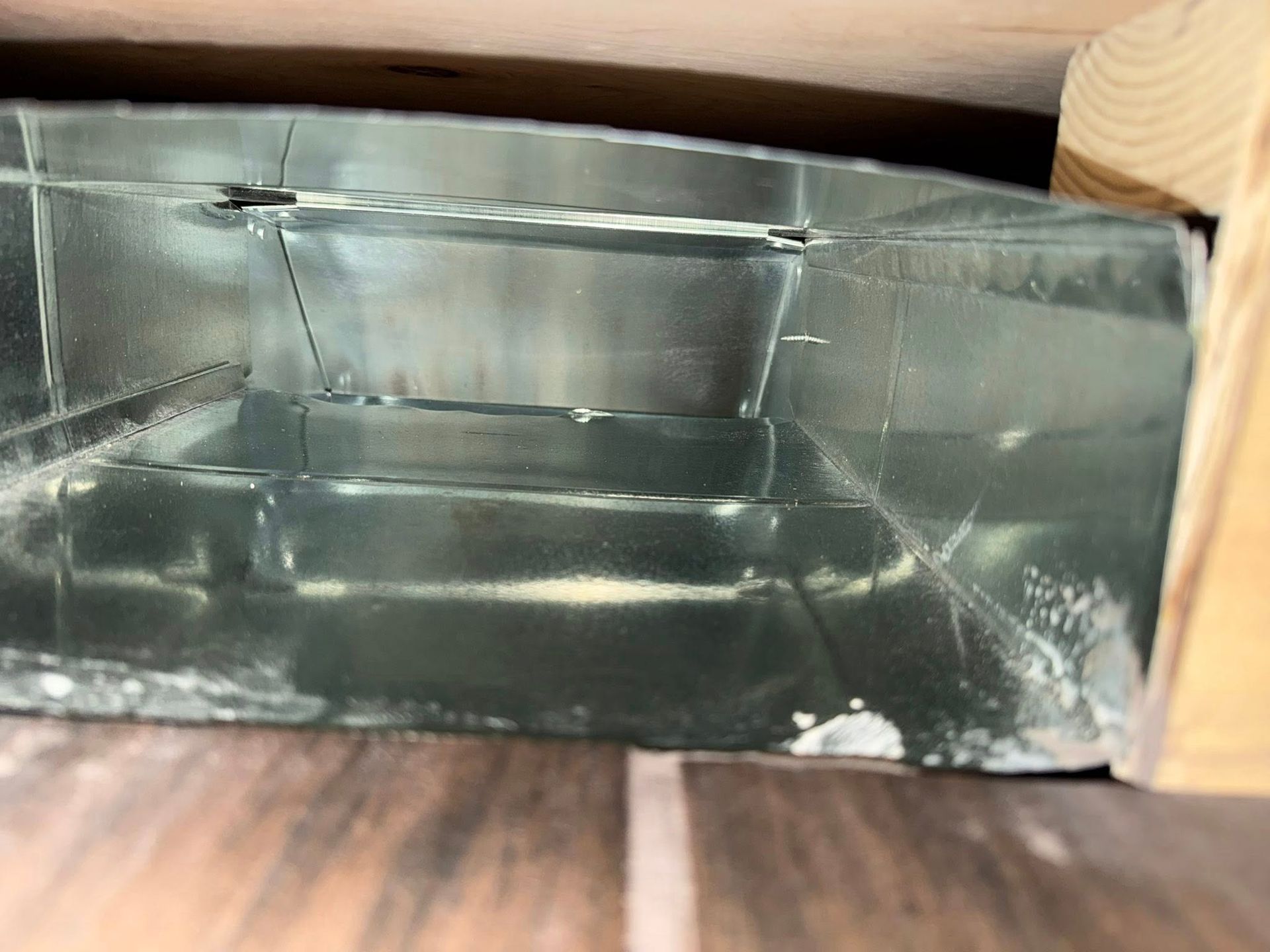 Inside a metal duct or vent, showing reflective silver interior panels and a wooden frame edge.