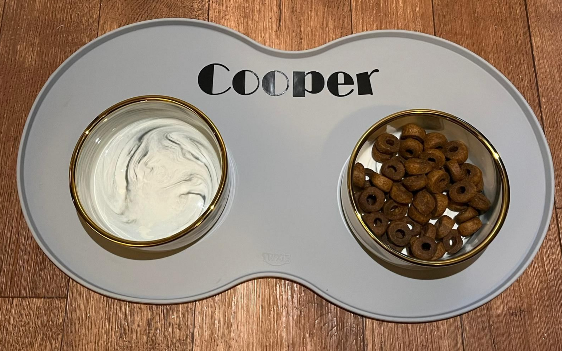 Hondenbakken op een grijze mat met Coopers naam erop. In de ene bak zit wit en grijs voer, in de andere bruine brokjes.