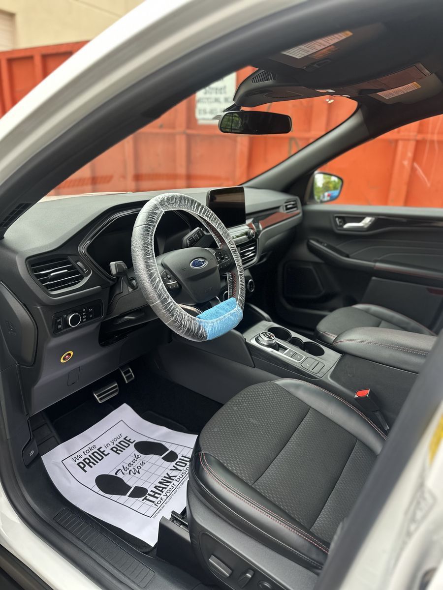 Interior of a white Ford Escape, black seats, steering wheel cover, paper floor mat, and empty mask hanging.
