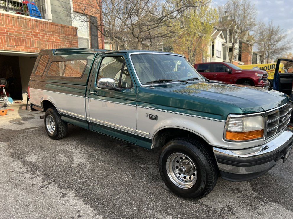Green and silver Ford F-150 pickup truck with a camper shell parked on asphalt in front of a brick building.