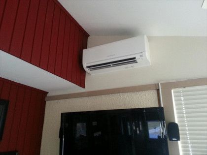 White air conditioning unit mounted on a beige wall above a TV, near a red wall and window blinds.