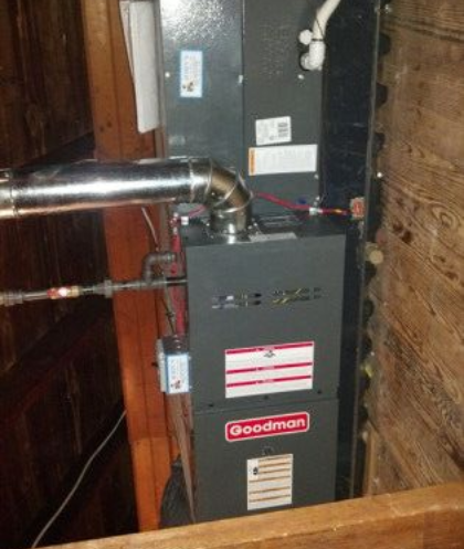 A Goodman furnace installed in a wooden enclosure. Silver exhaust pipe, gas line, and control box visible.