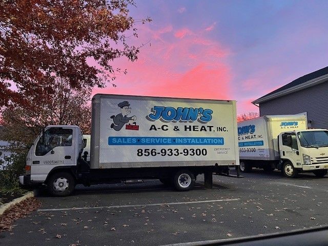 Two John’s A-C & Heat trucks parked outside at dusk with pink sky and autumn trees