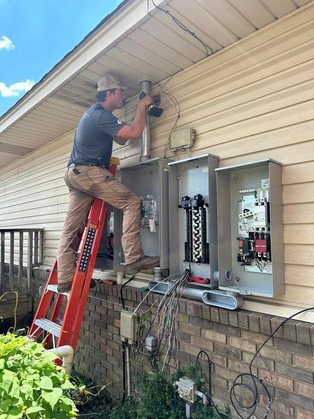 A man is standing on a ladder working on an electrical box on the side of a house.