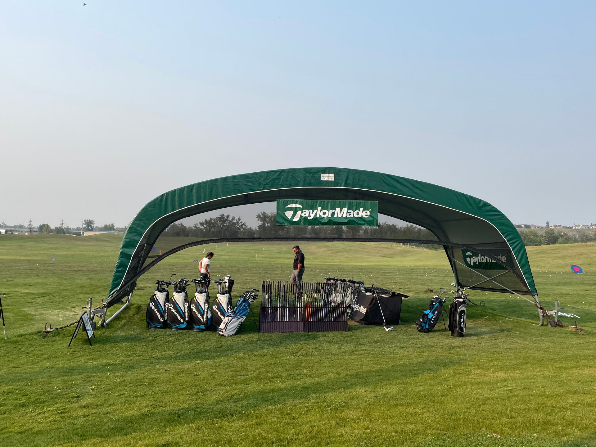 TaylorMade golf equipment display under a green canopy on a grassy field.