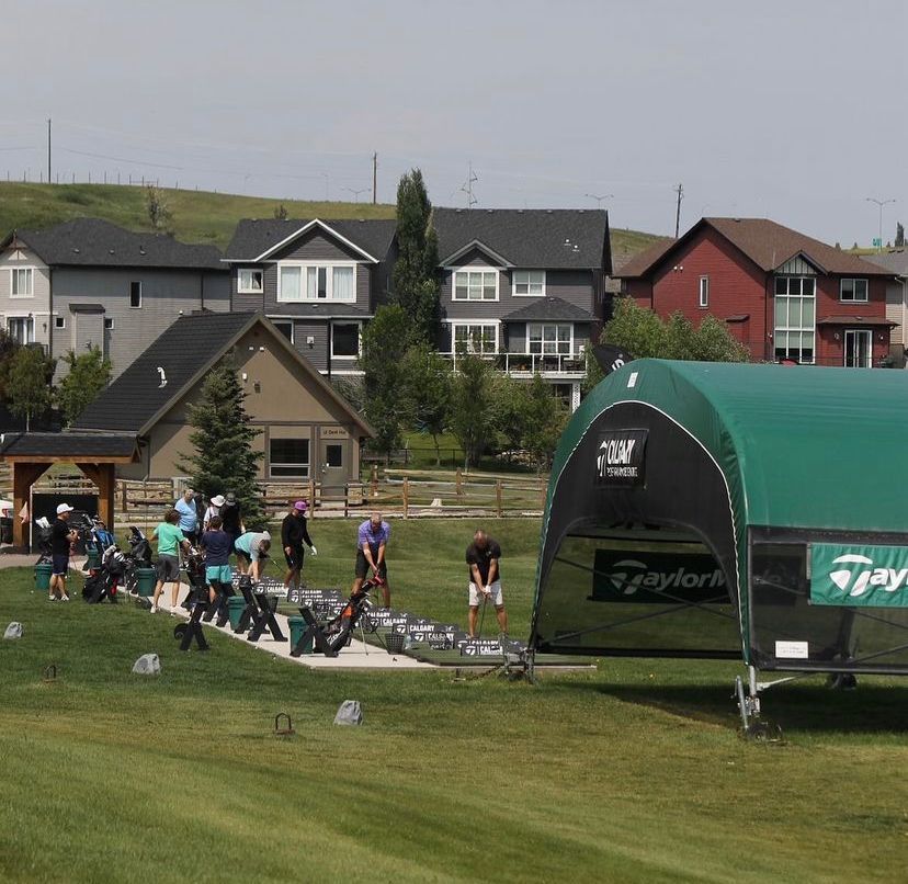 Golfers practice on a green field. A green tent with TaylorMade logo is in the foreground. Houses in the background.