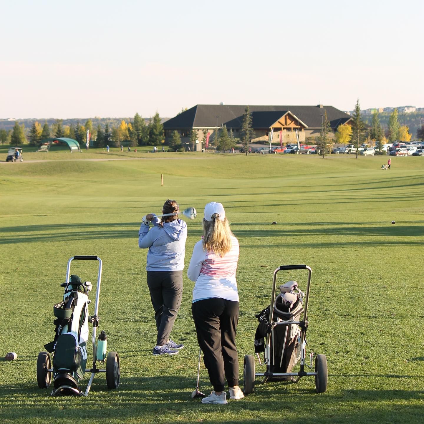 Two people golfing on a green course, with golf carts. A clubhouse and trees are in the background.