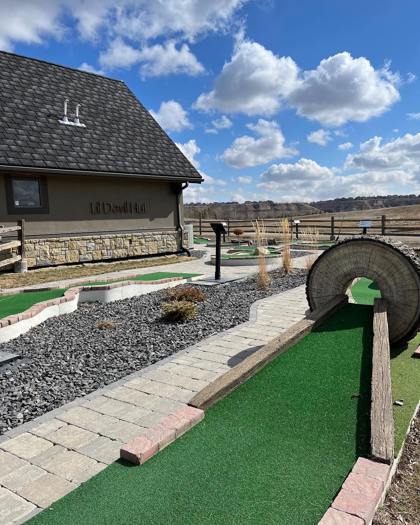 Miniature golf course with green turf, brick borders, and a stone arch, set against a building and open field.
