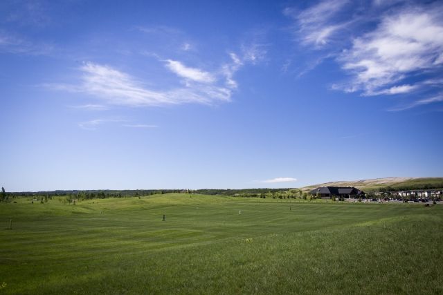 Vast green field under a bright blue sky with scattered clouds, a building is visible in the distance.