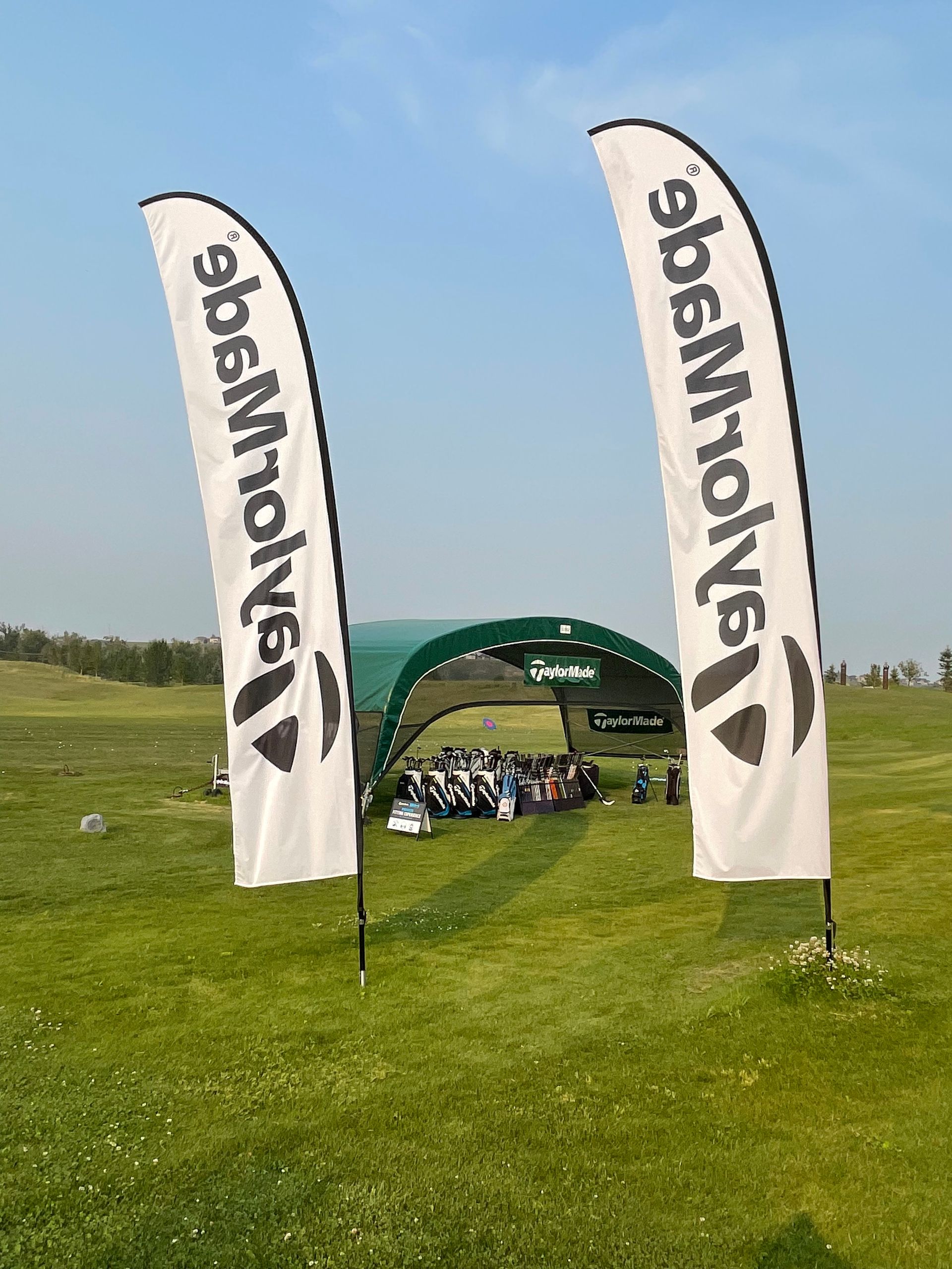Two white TaylorMade flags flank a green tent on a golf course under a blue sky.