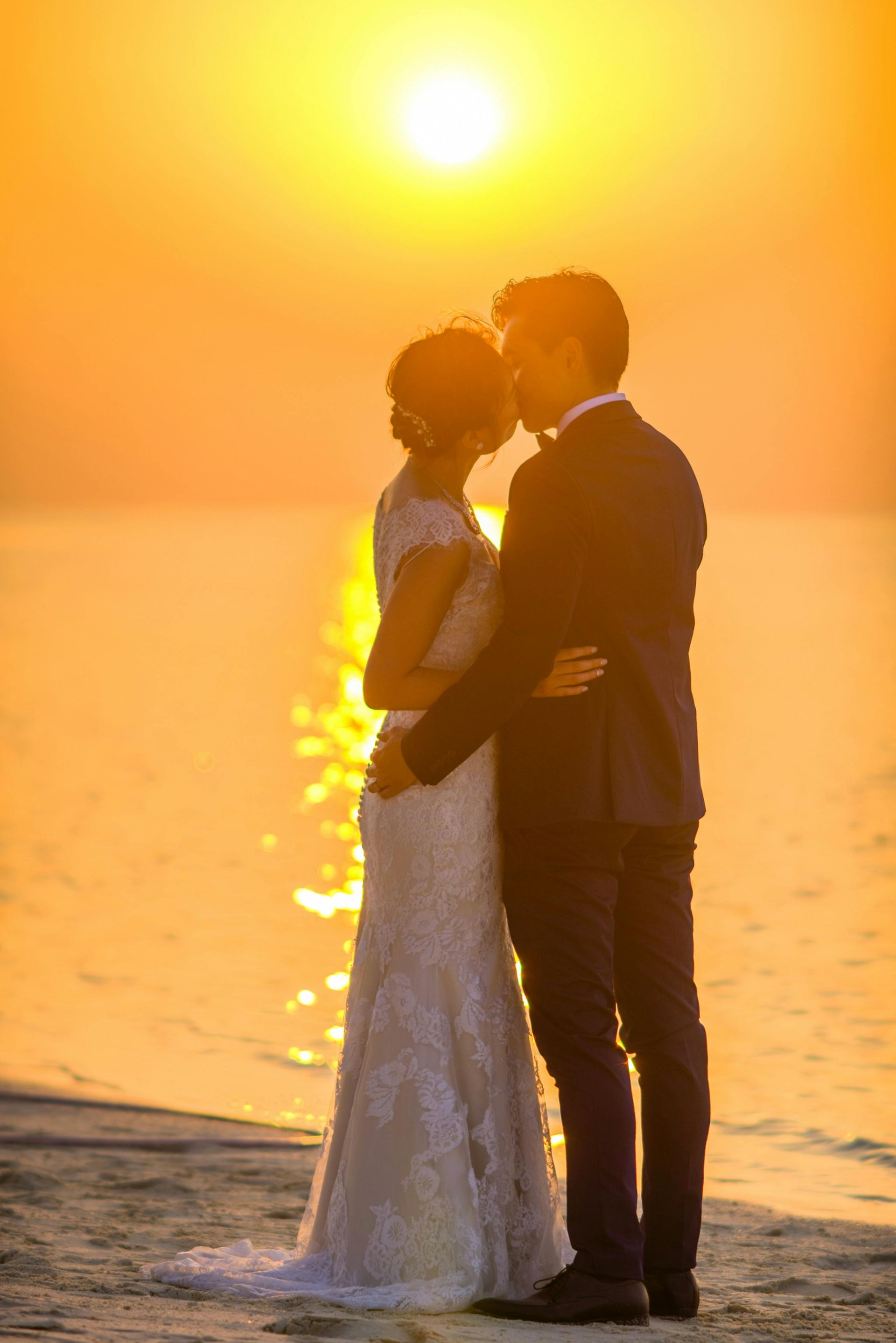 A bride and groom are kissing on the beach at sunset.