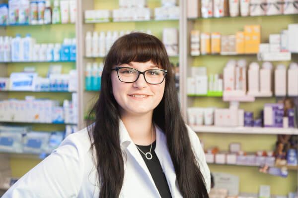 Eine Apothekerin mit Brille steht vor einer Apotheke.