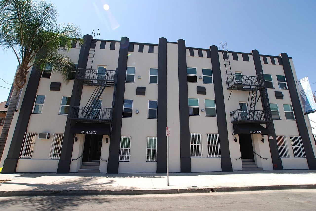 Three-story beige apartment building with dark trim, fire escapes, and many windows on a sunny day.
