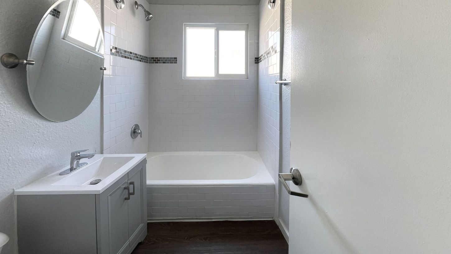 Small bathroom with a white tub, sink, and walls; dark floor, a window, and a silver mirror.