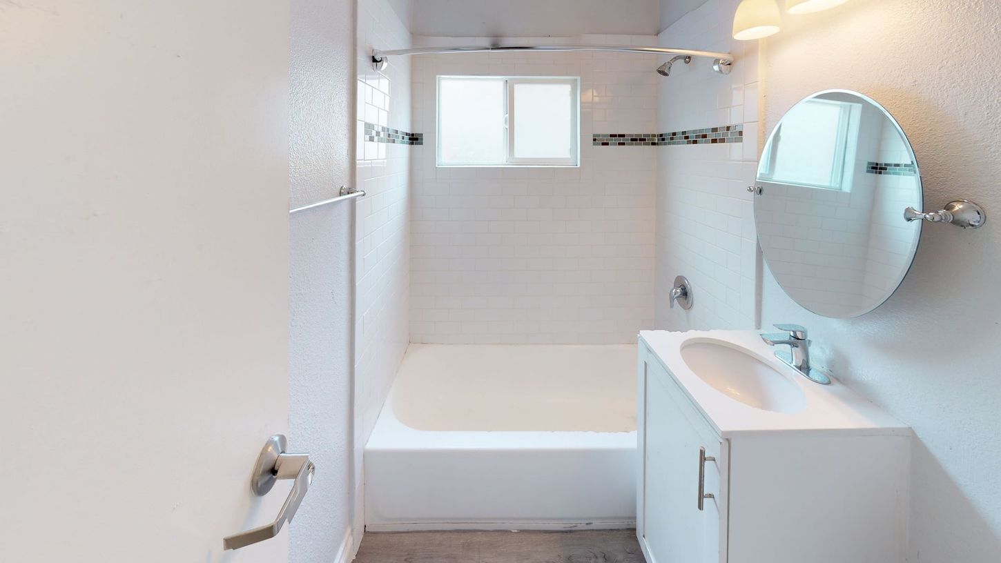 A small, white bathroom with a tub, sink, mirror, and a small window.