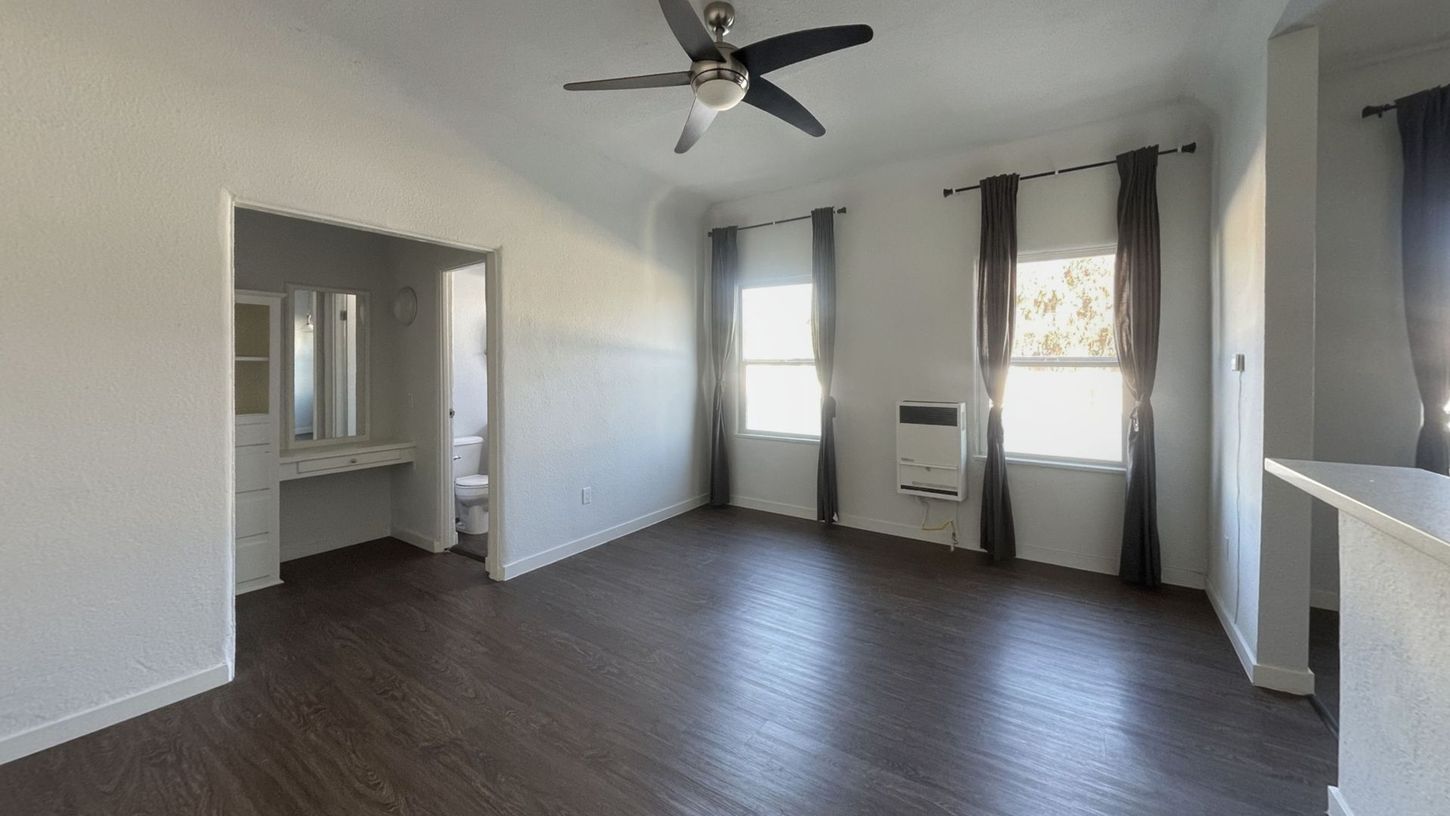 Empty apartment interior with dark wood floors, white walls, two windows with curtains, and a ceiling fan.