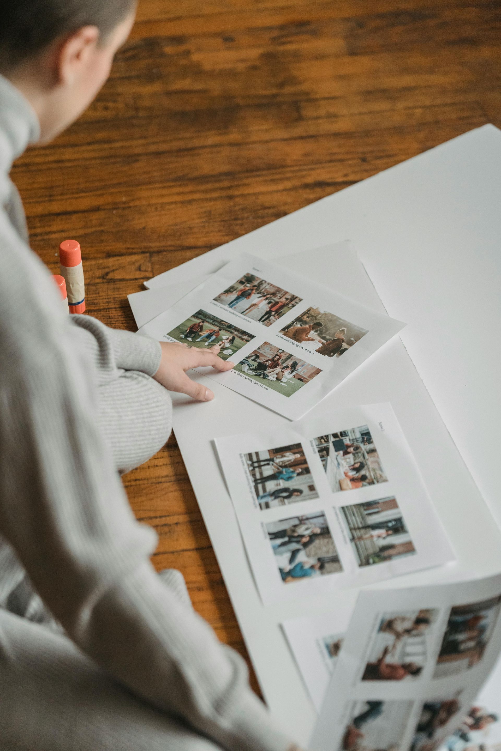 A person sits on a wood floor, organizing printed photo collages on large white sheets of paper.
