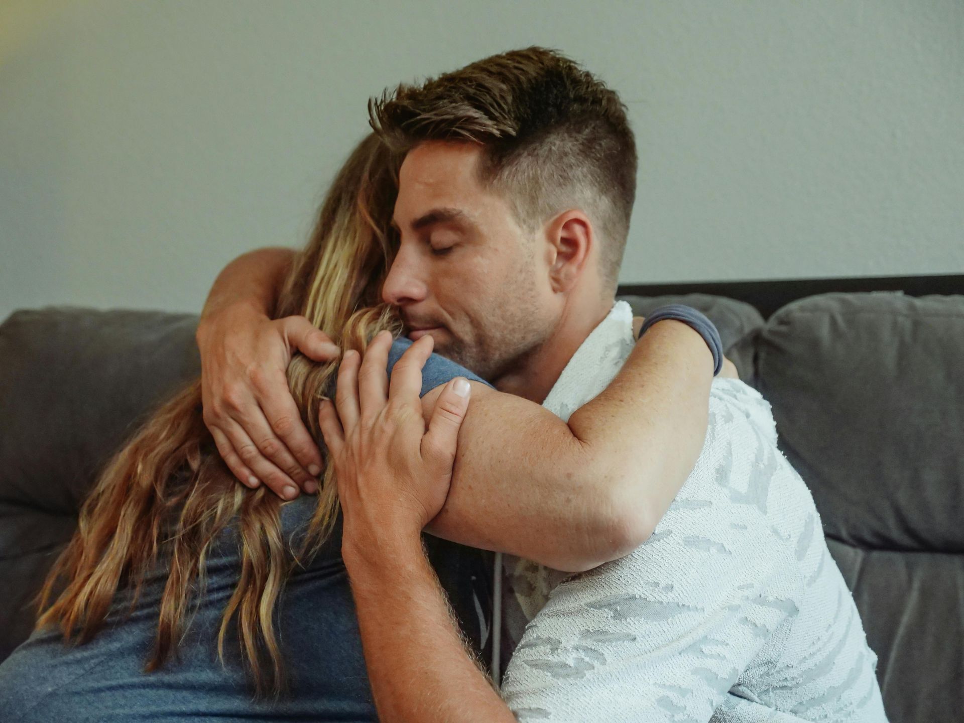A man wearing a white patterned shirt embraces a person with long hair while sitting on a gray couch.