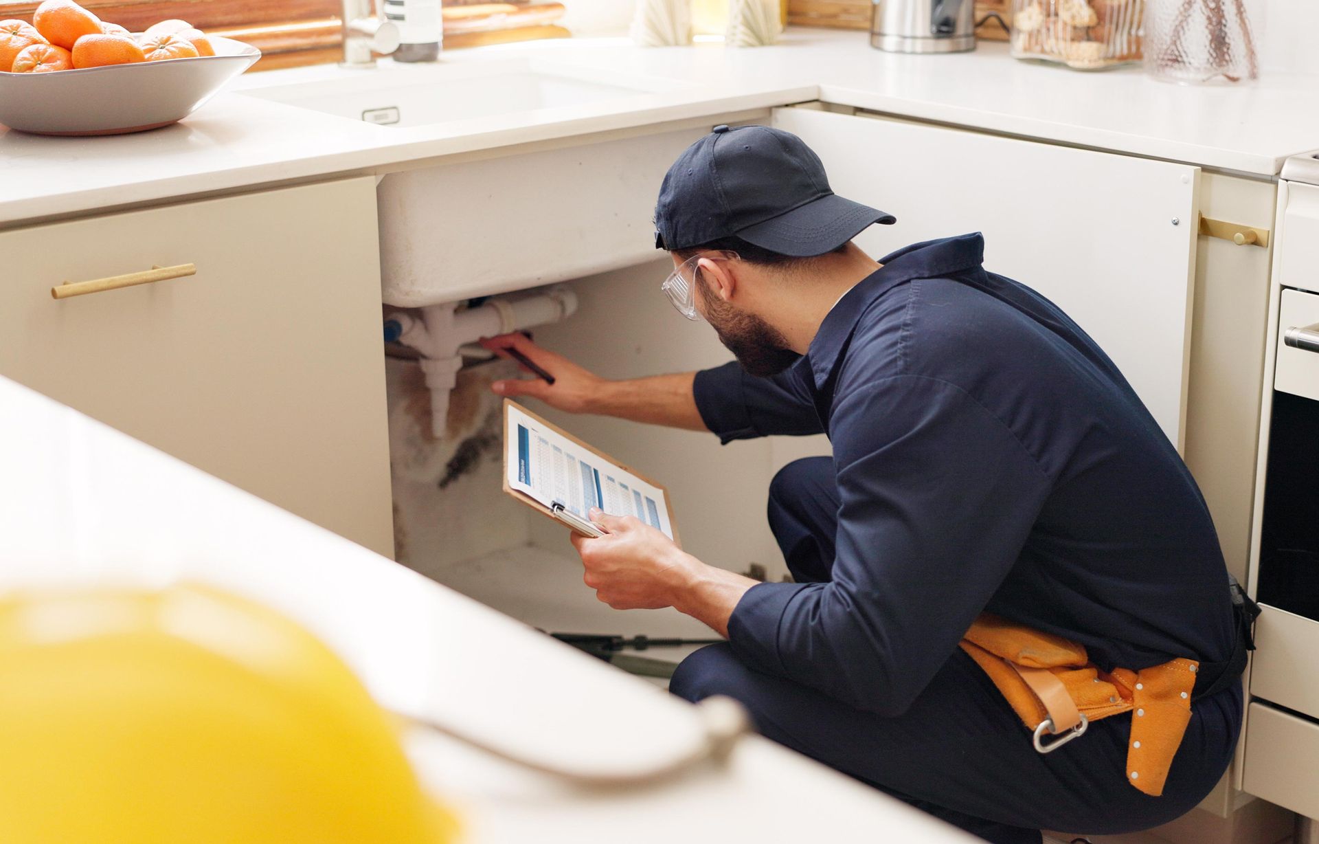 Plumber inspecting kitchen sink pipes, holding clipboard, wearing dark blue uniform and cap.