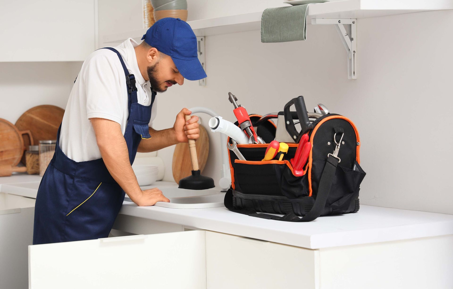 Plumber in blue uniform, using plunger, with toolbox on white kitchen counter.